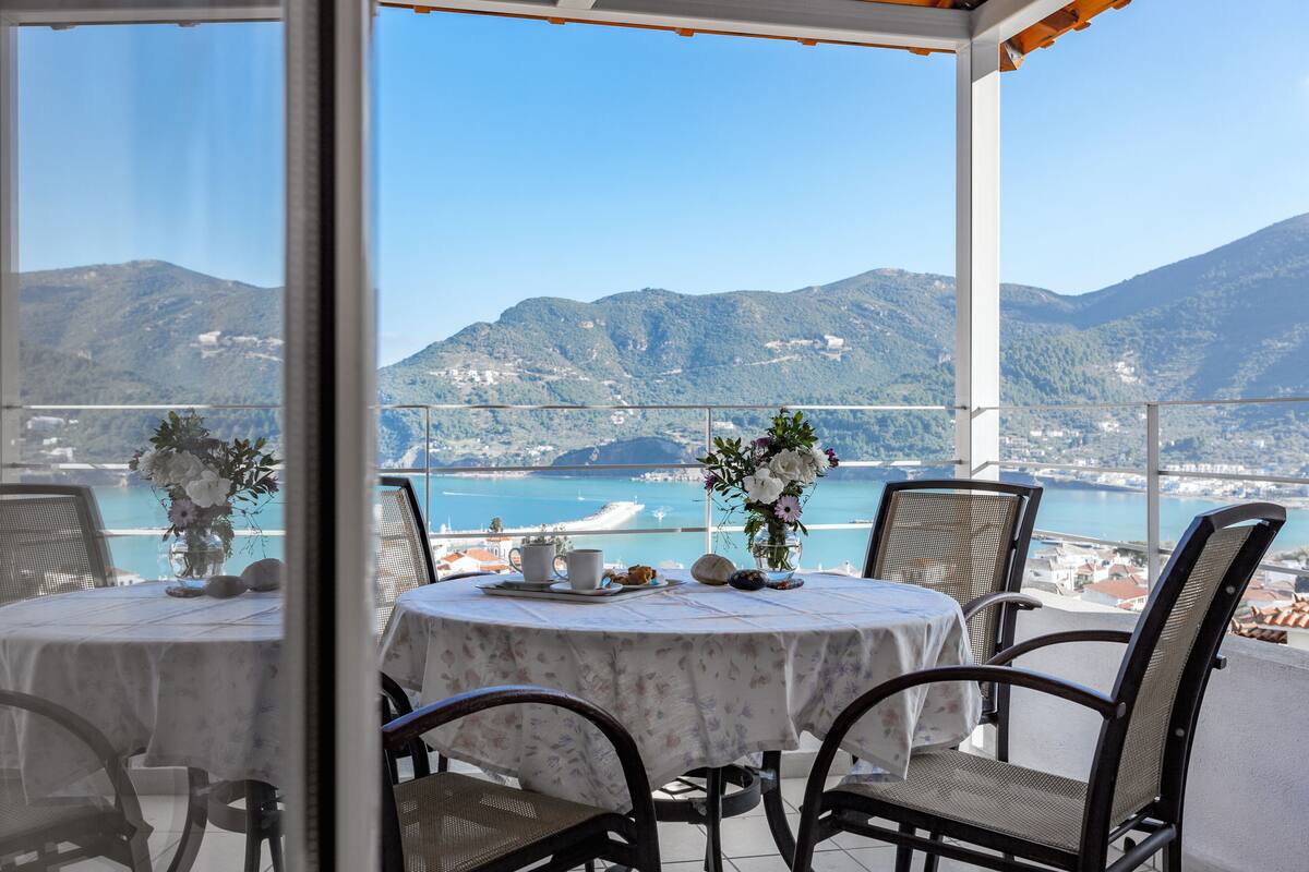 Une terrasse avec des tables et des chaises rondes, un vase rempli de fleurs et une vue sur une zone côtière, avec des montagnes en arrière-plan.