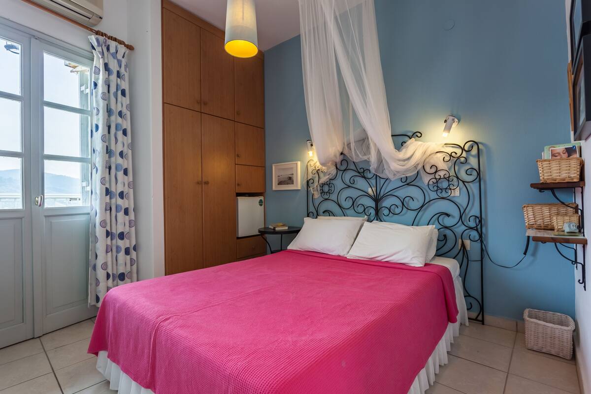 Une chambre à coucher comprenant un lit, une table de chevet, une étagère fixée au mur, une fenêtre avec des rideaux et un ventilateur de plafond.