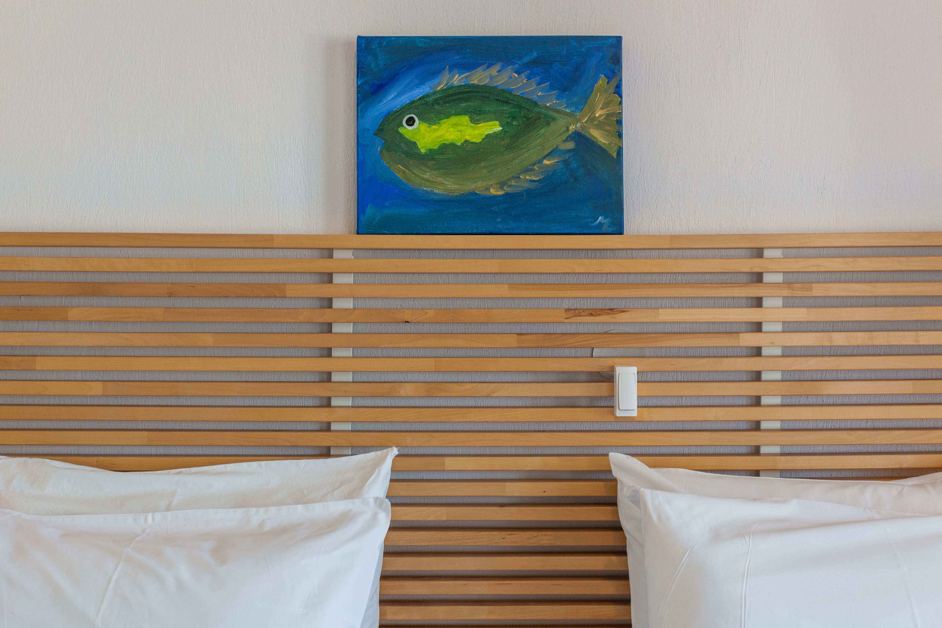 Ett rum med en sänggavel av träribbor, två vita kuddar och en tavla med ett fiskmotiv på väggen.
