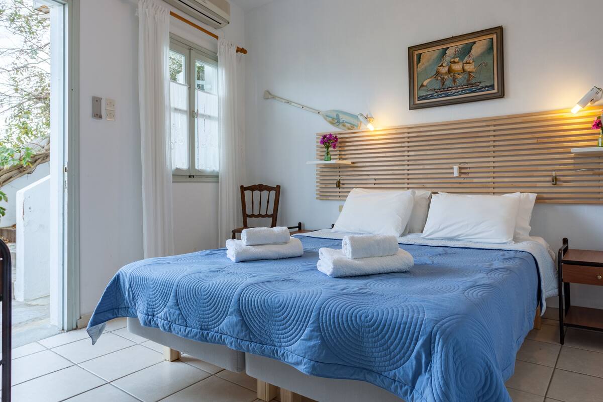 Une chambre à coucher avec un lit, des oreillers blancs, une couverture bleue, une tête de lit en bois, une table de chevet et un tableau représentant un navire.