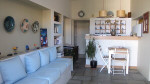 Una sala de estar con un sofá azul claro, una mesita blanca pequeña y una cocina con mostrador y lámparas colgantes.
