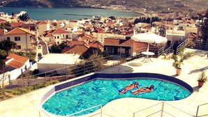 Una piscina con dos personas flotando, rodeada de un suelo de baldosas y vistas a un pueblo costero.