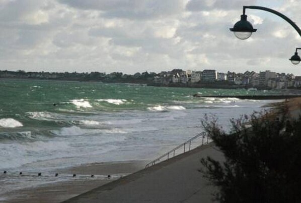Vista costera con un paseo pavimentado a lo largo de la playa, un poste de luz y una línea de costa distante con edificios.