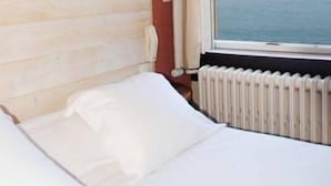 Habitación de hotel con cabecera de madera, ventana con vista al mar y cama blanca.