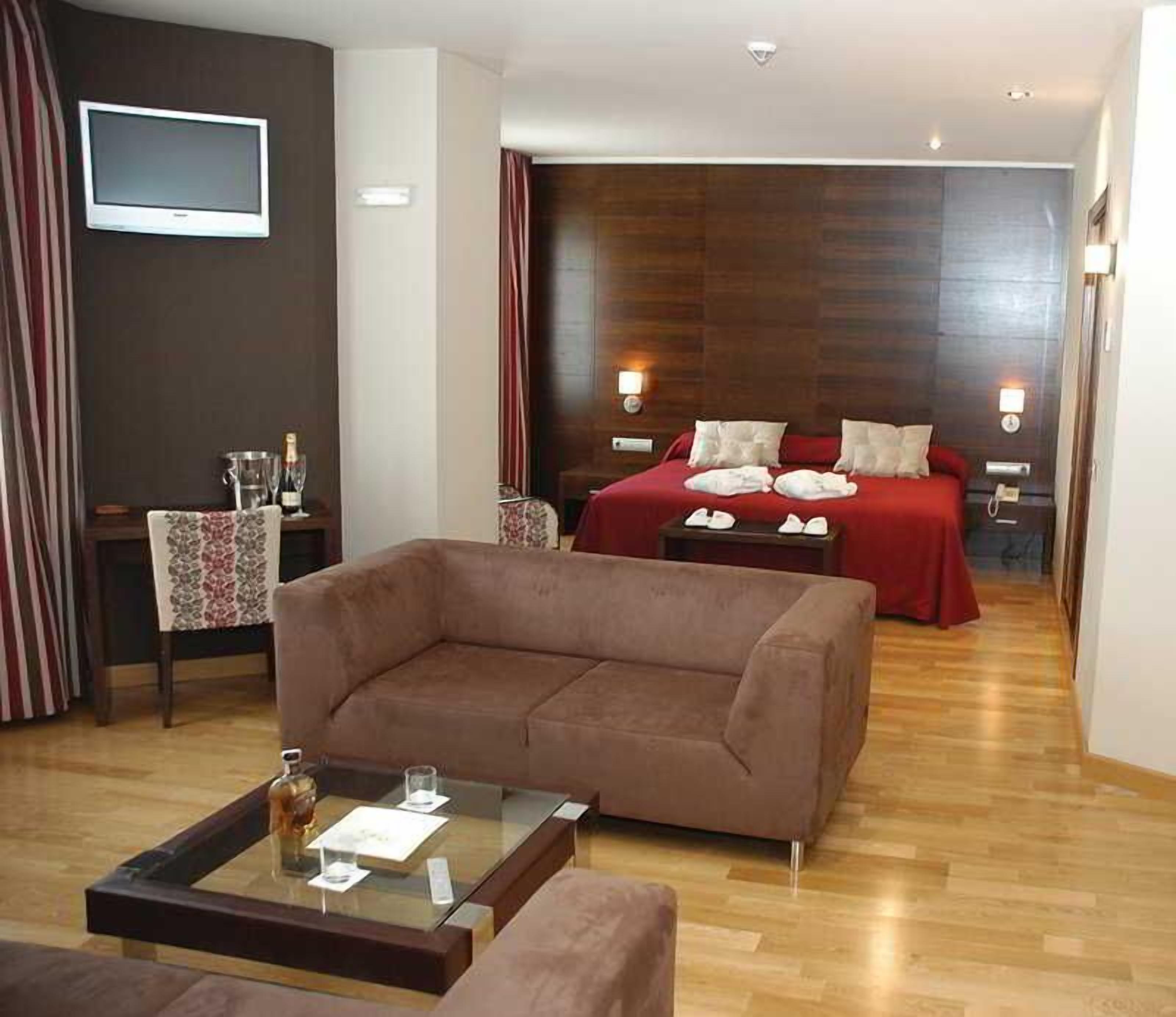 Habitación de hotel con sofá marrón, una mesa de centro de cristal, cama con ropa de cama roja y cabecera de madera.