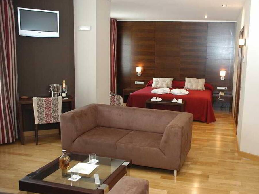 Habitación de hotel con sofá marrón, una mesa de centro de cristal, cama con ropa de cama roja y cabecera de madera.