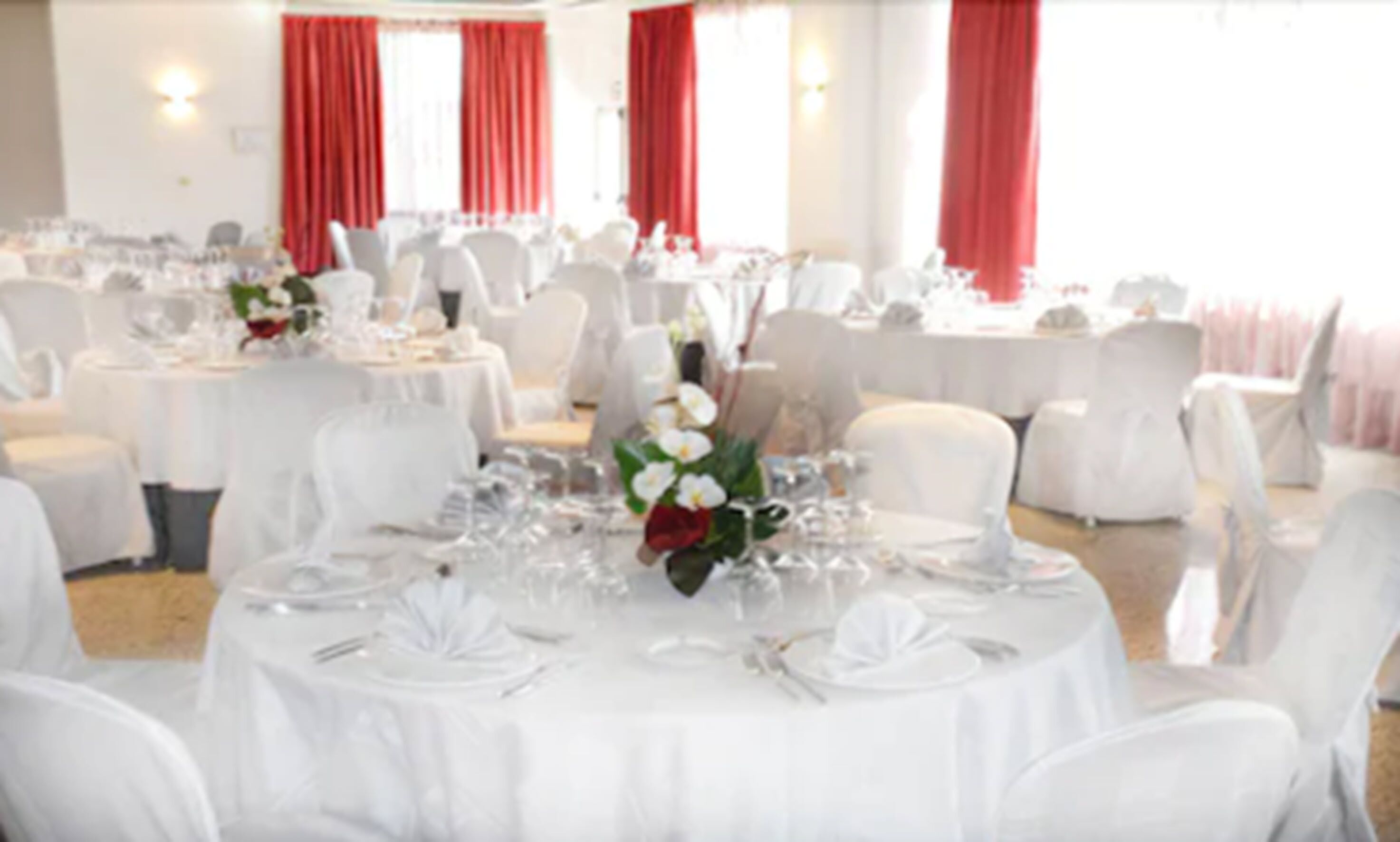 A banquet hall with round tables set for dining, white chairs, and red curtains.