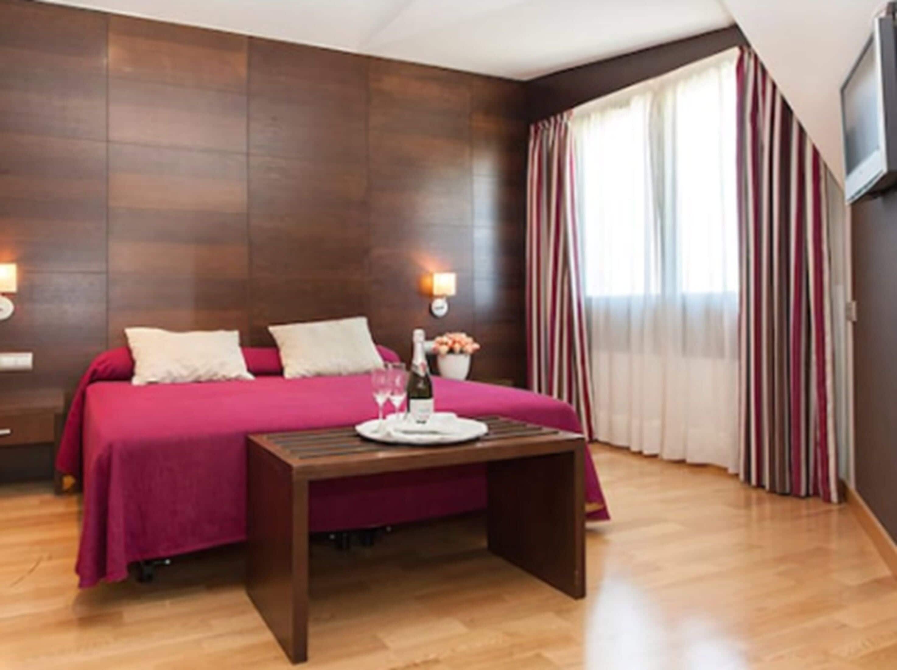 A hotel room with a bed, bedside tables, a nightstand, a wooden bench, a bottle of champagne, glasses, and a vase with flowers.