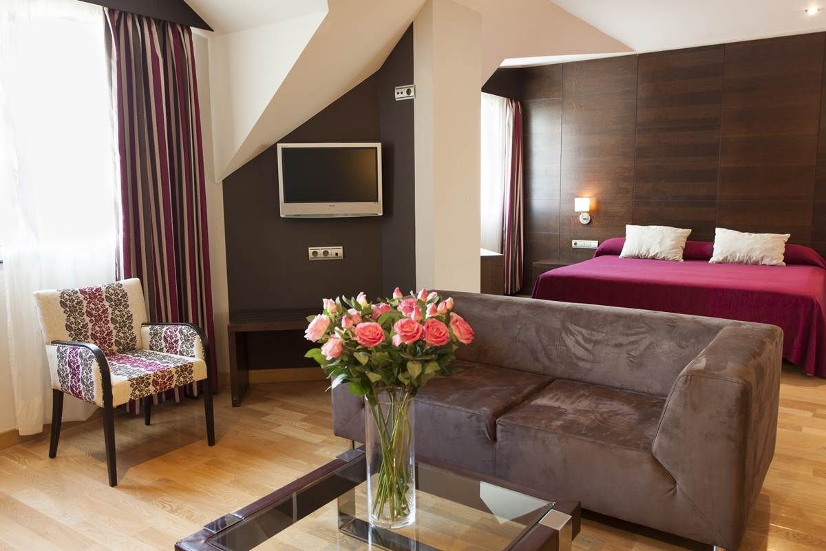 A modern hotel room with a sofa, a chair, a TV mounted on the wall, and a bed with a red bedspread.