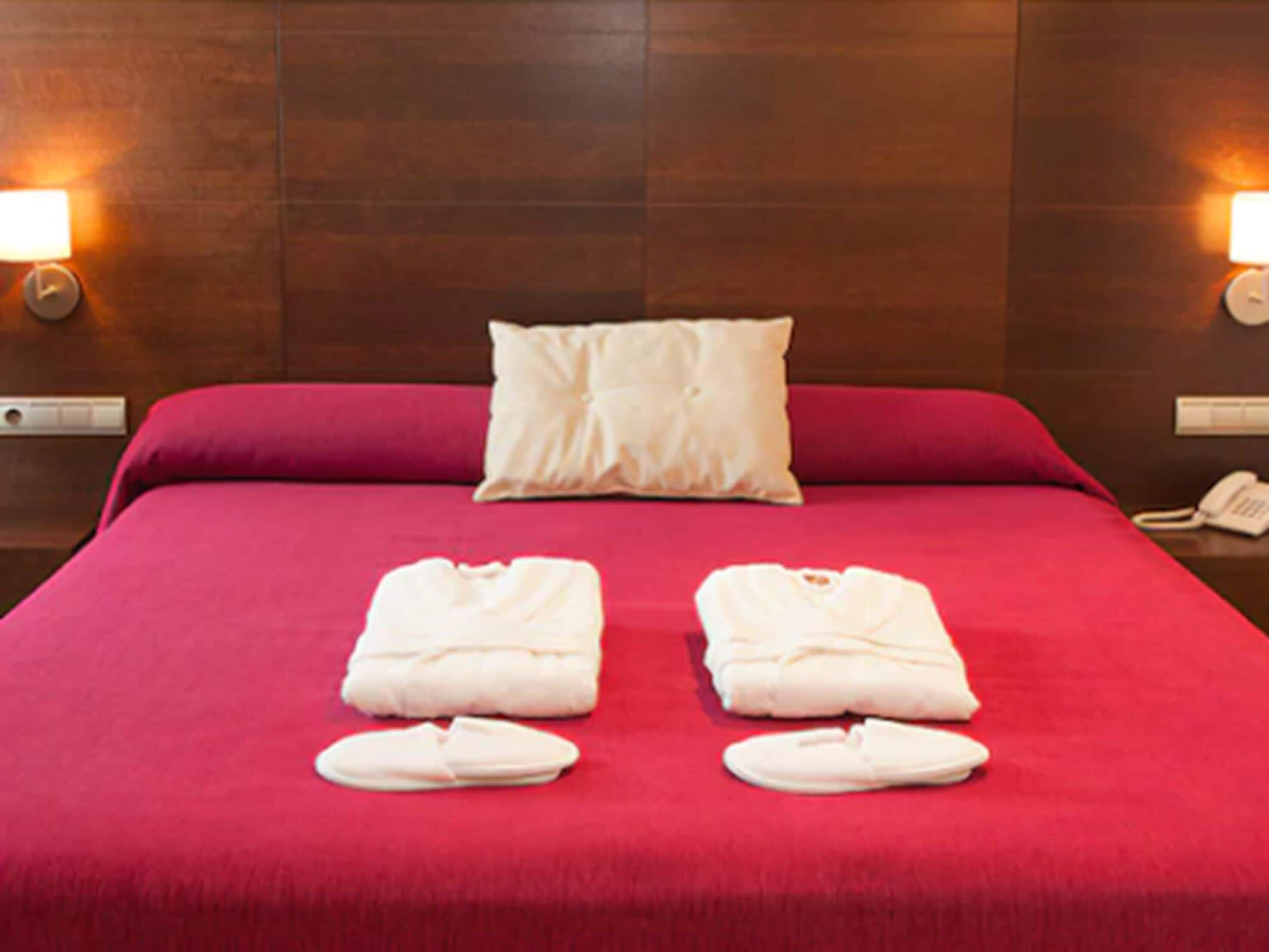 Camera d'albergo con copripiumino rosso, due cuscini bianchi e un asciugamano piegato e ciabatte bianche sul letto.