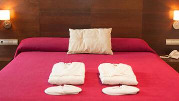 Camera d'albergo con copripiumino rosso, due cuscini bianchi e un asciugamano piegato e ciabatte bianche sul letto.