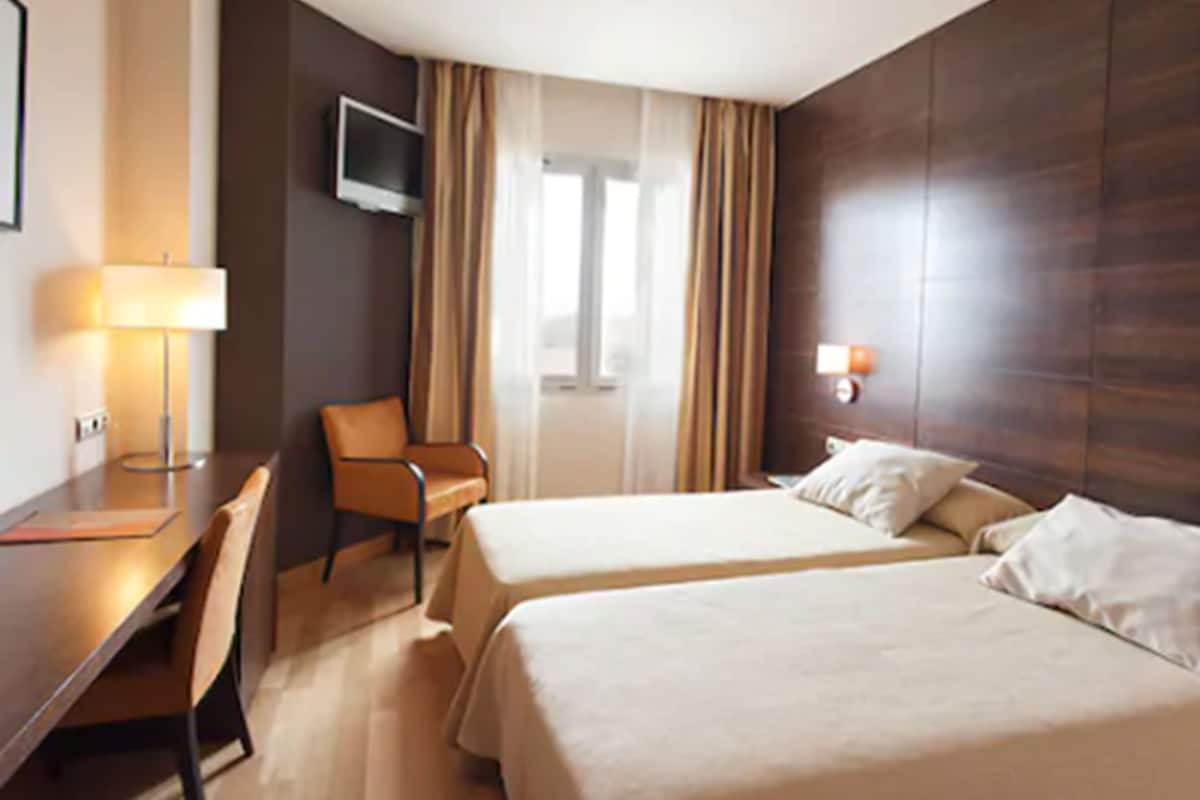 Une chambre d’hôtel avec deux lits, un bureau, une chaise, un téléviseur fixé au mur et une fenêtre avec des rideaux.