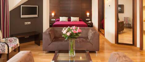A modern hotel room with a large bed, a sofa, a coffee table with flowers, and a TV on the wall.