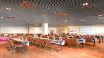 A large conference room with rows of tables and chairs, a high ceiling with a grid pattern, and a projector screen at the front.