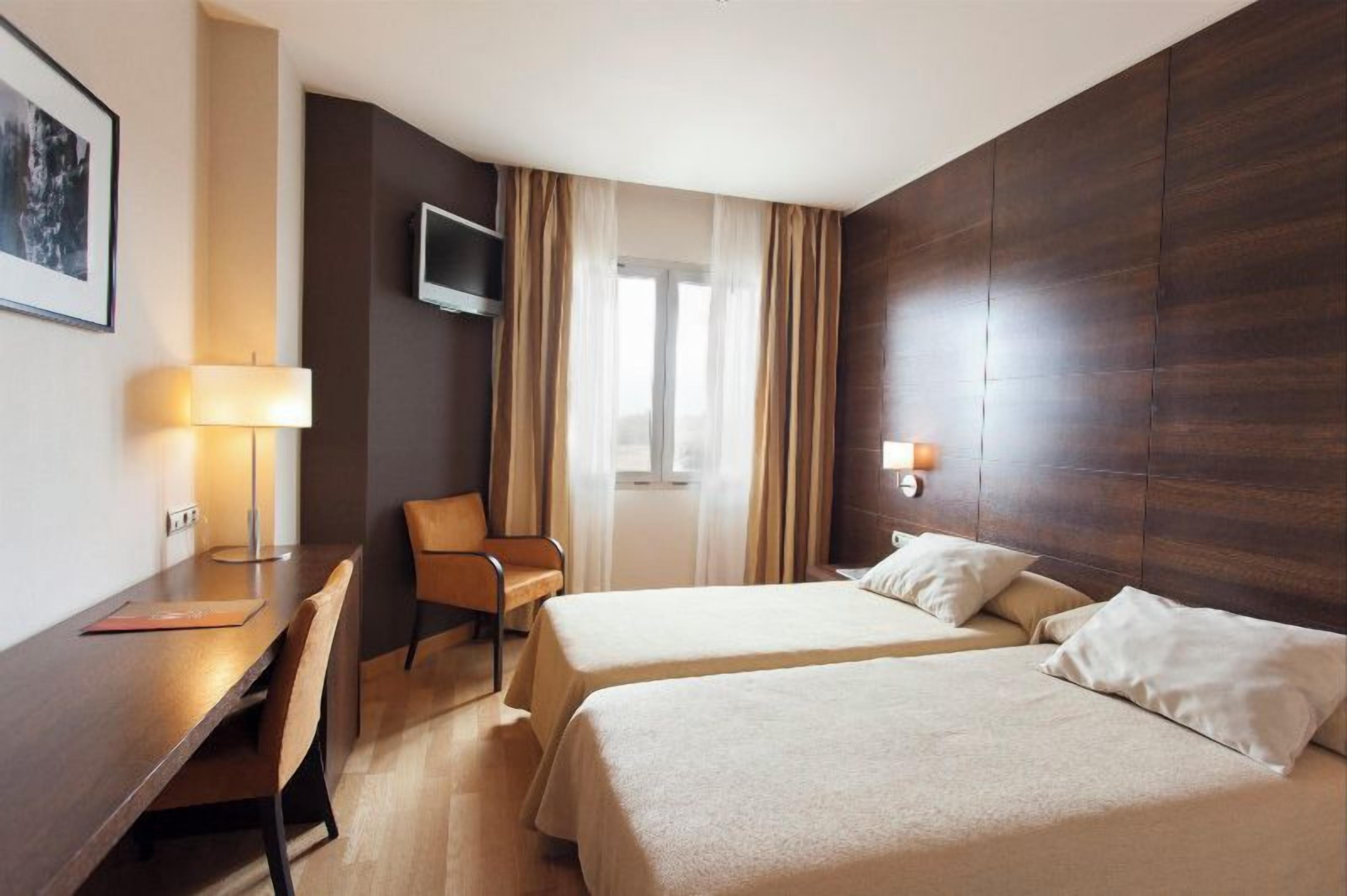 A hotel room with two beds, a desk, a chair, a TV mounted on the wall, and a window with curtains.