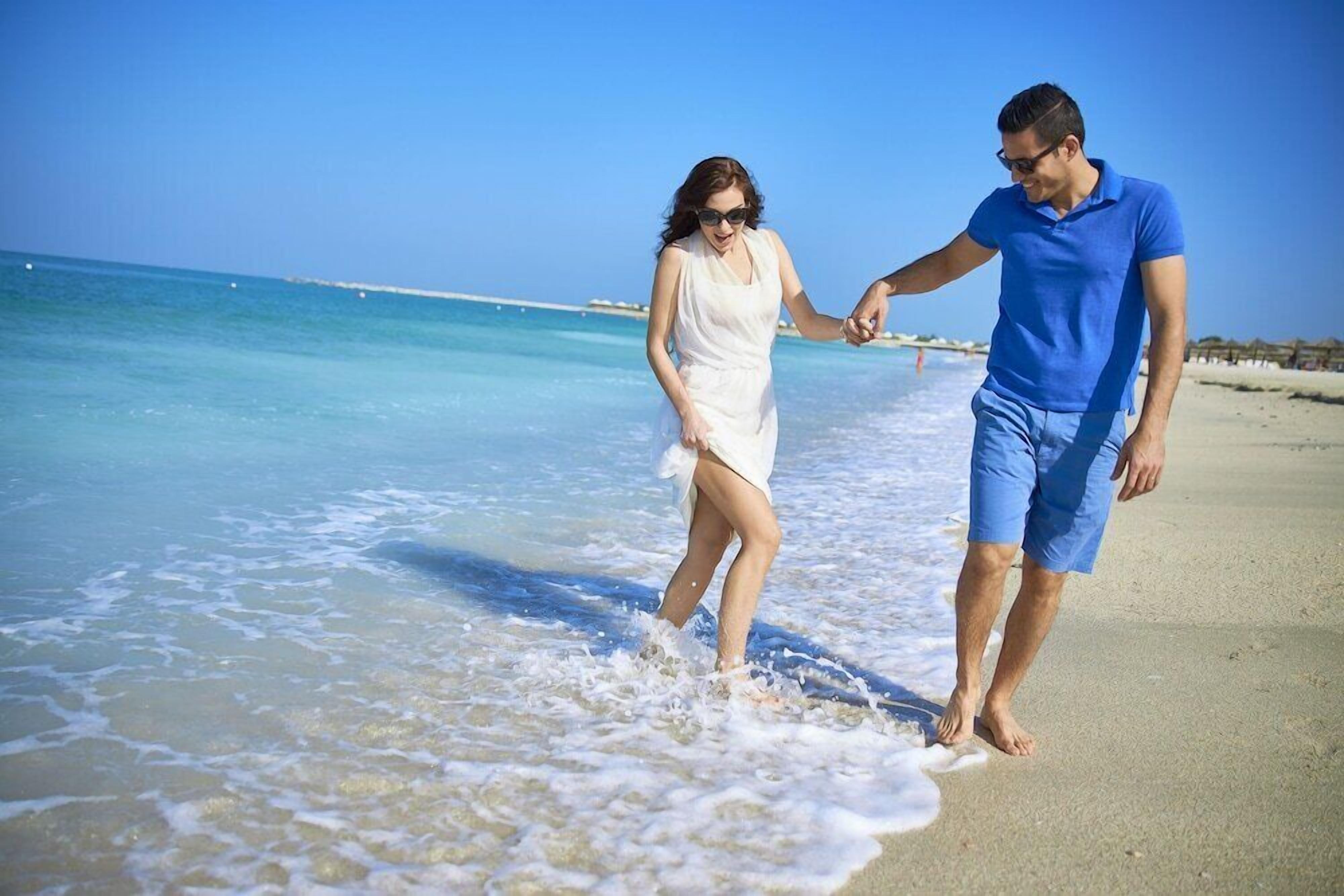 A couple walking hand in hand on a beach with clear blue water and a sandy shore.