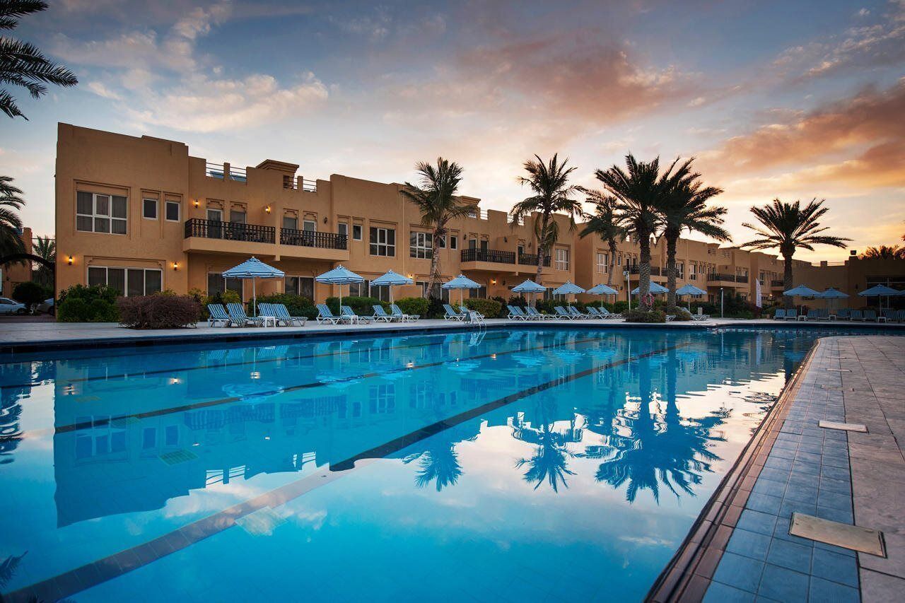 A swimming pool with lounge chairs and umbrellas, surrounded by palm trees and a building with balconies.