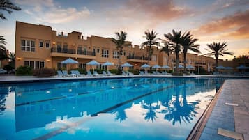 Una piscina con sillones de descanso y sombrillas, rodeada de palmeras y un edificio con balcones.