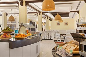 Zona de buffet con diferentes opciones de comida, entre ellas frutas y pan, además de un área de descanso con sillas blancas.