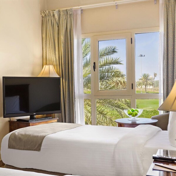 A hotel room with a bed, a television, a window with curtains, and a view of palm trees.