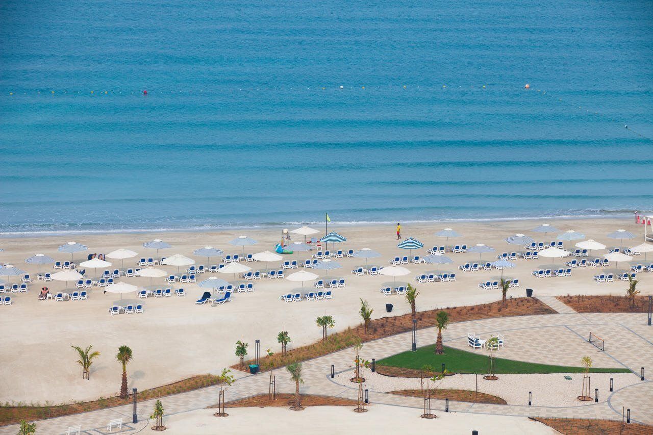 Een strand met rijen ligbedden en parasols, een vlag en palmbomen.