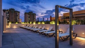 Une terrasse sur le toit, dotée de chaises longues et d’une structure métallique, offrant une vue sur le paysage urbain au crépuscule.
