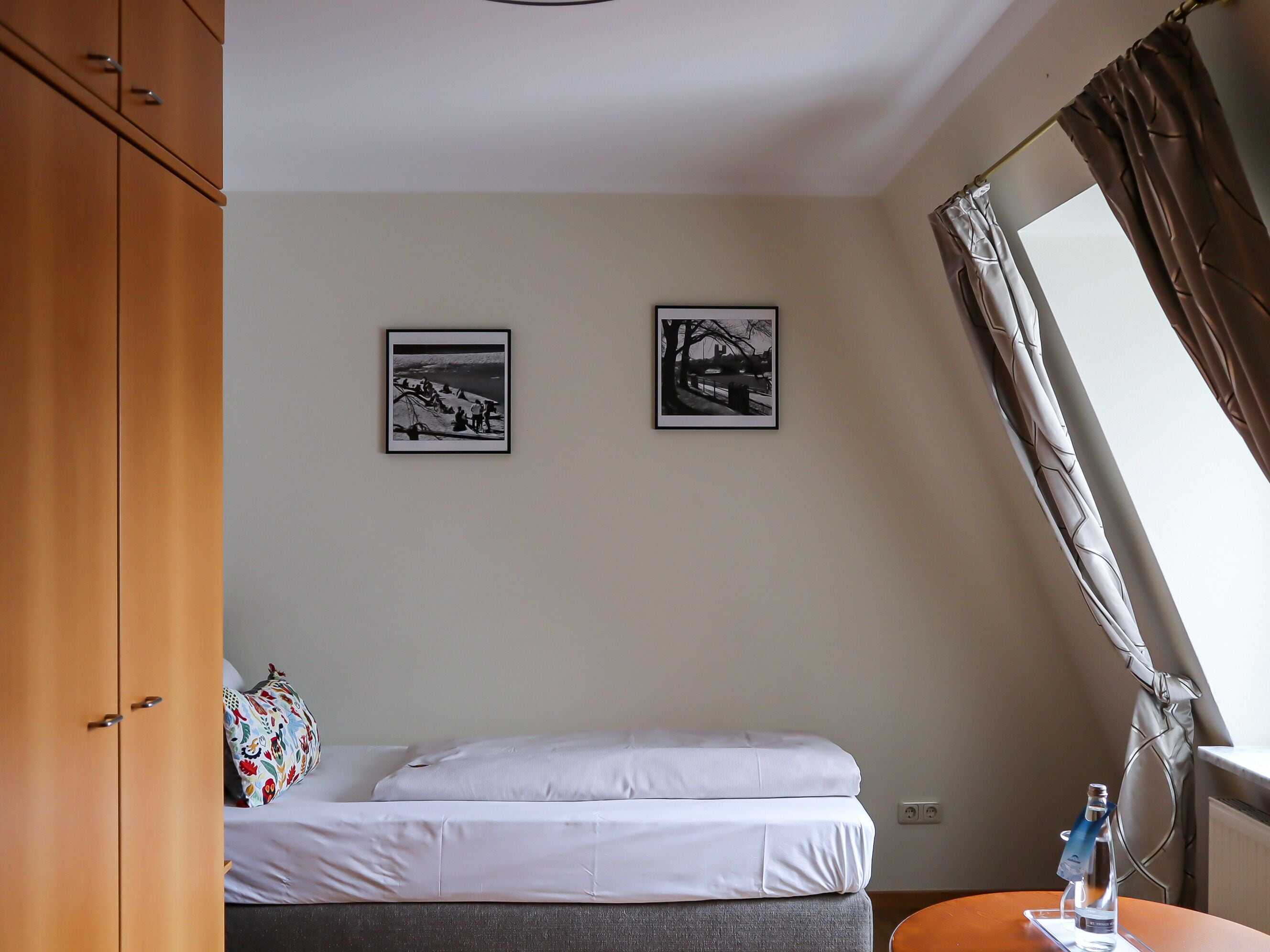 A bedroom with a bed, two framed pictures on the wall, a window with curtains, and a wooden wardrobe.