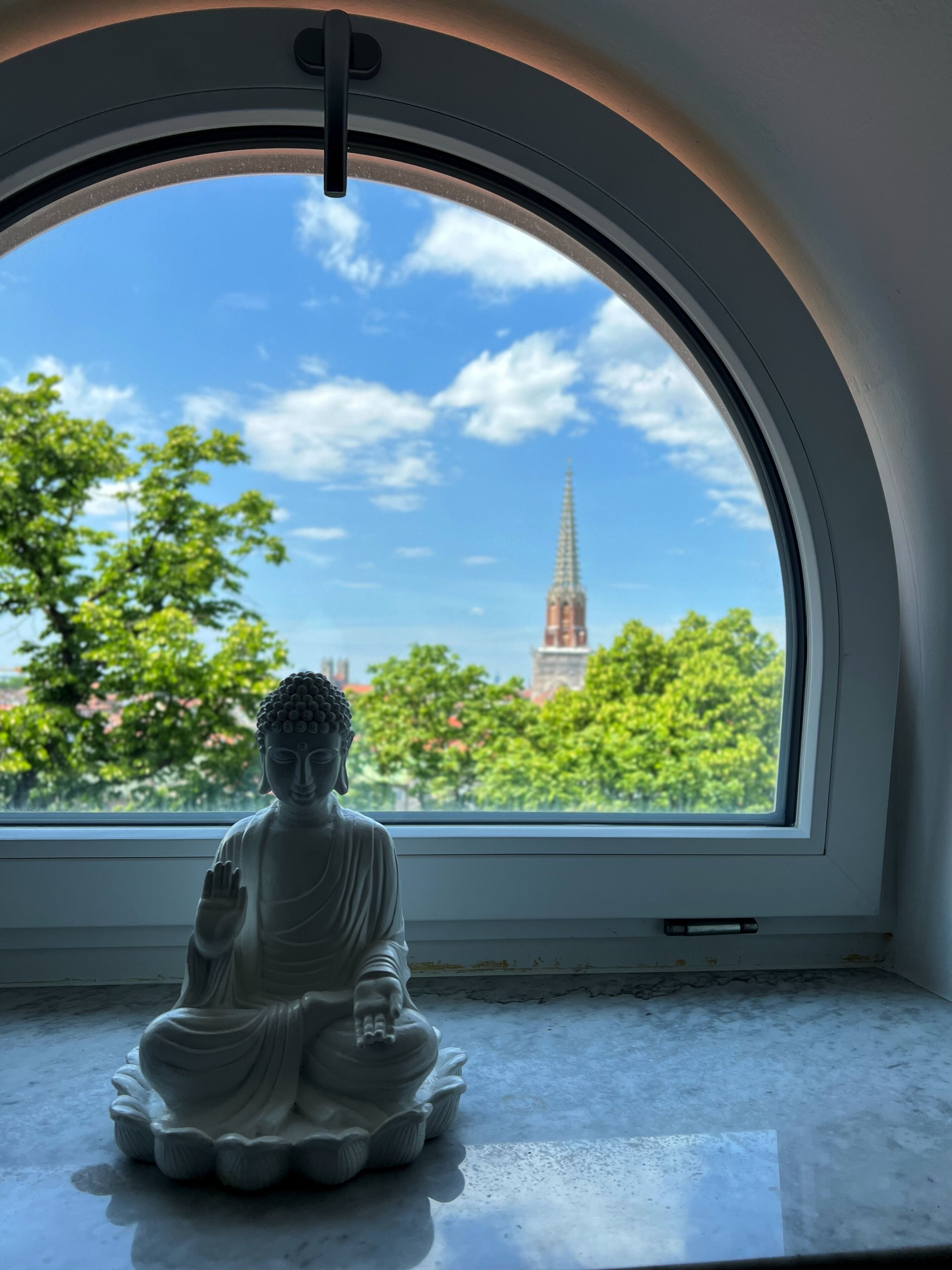 Boeddhabeeld in meditatieve houding op een vensterbank, met uitzicht op een kerktoren en bomen.