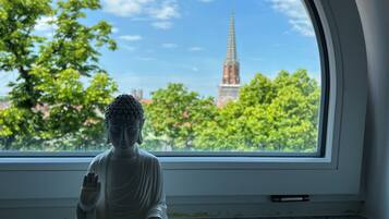 Boeddhabeeld in meditatieve houding op een vensterbank, met uitzicht op een kerktoren en bomen.