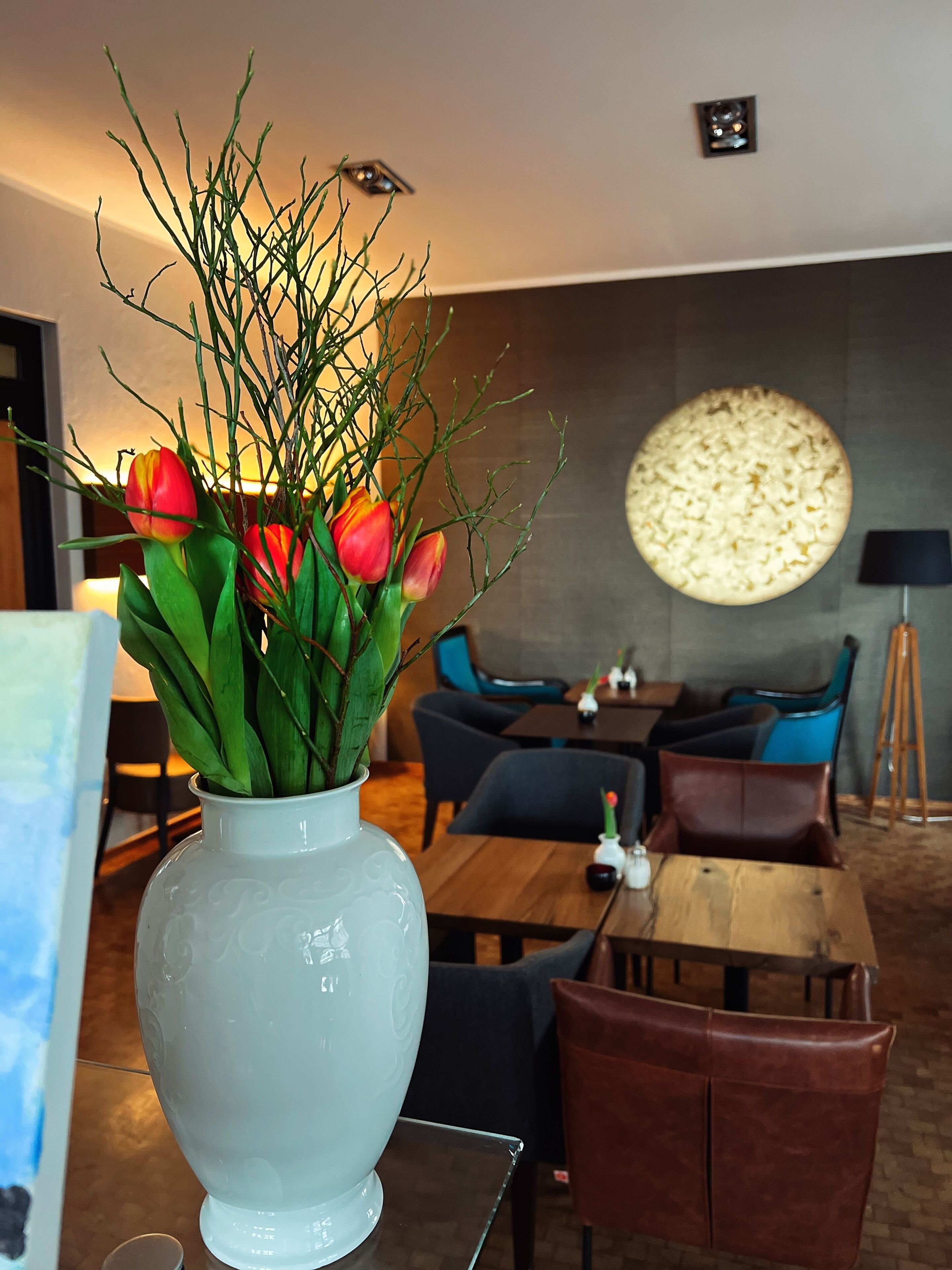 Un interior moderno con un jarrón blanco de tulipanes sobre una mesa, un adorno circular en la pared y un comedor con sillas y una mesa.