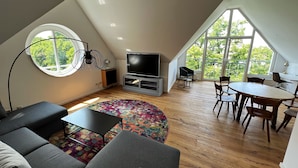 A modern loft apartment with a large skylight, a cozy seating area with a sofa and coffee table, a dining table with chairs, and a flat-screen TV on a stand. - Hotel Prinz (Munich)