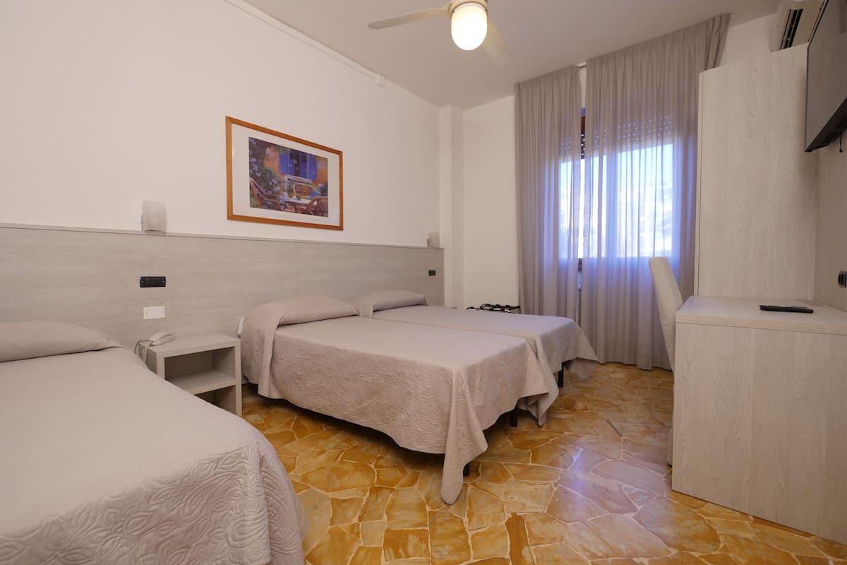 A hotel room with two beds, a desk, a chair, a ceiling fan, a framed picture on the wall, and a window with curtains.