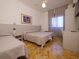 Una camera d'albergo con due letti, una scrivania, una sedia, un ventilatore a soffitto, un quadro appeso al muro e una finestra con tende.