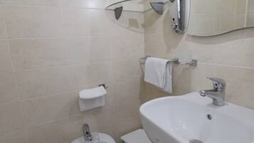 Un baño con lavamanos blanco, bidé e inodoro. Hay un estante de vidrio sobre el lavamanos y un perchero en la pared.