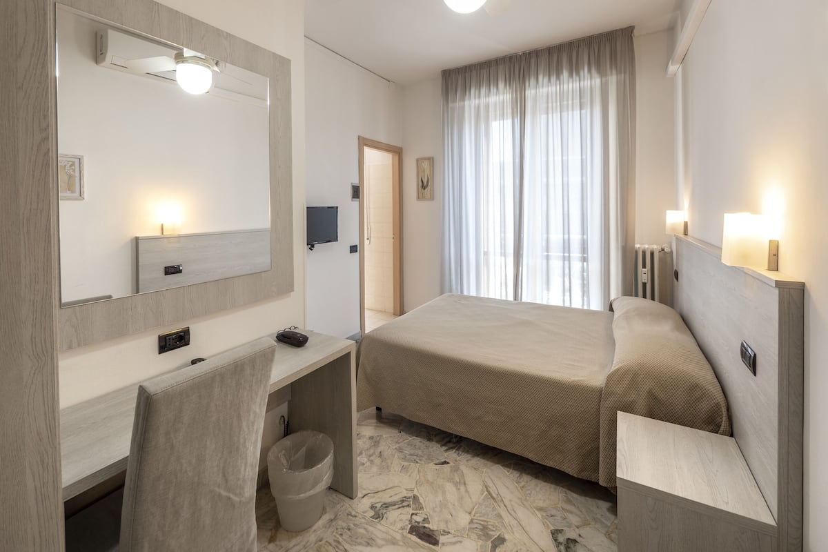A hotel room with a bed, a desk with a chair, a mirror, a television, and a window with curtains.