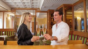 A couple toasting with champagne glasses in a restaurant with wooden furniture and a decorative ceiling. - Greif Hotel & Spa Trieste  (Trieste)