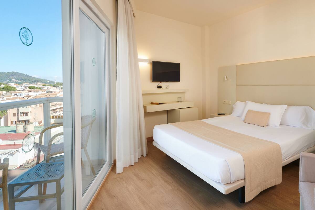 A hotel room with a bed, a TV, a desk, and a balcony with a view of the city.