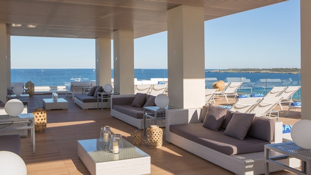A sunlit outdoor lounge area with wicker furniture, a wooden deck, and a view of the sea.