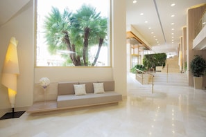A modern hotel lobby with a large window, a sofa with cushions, a staircase, and a potted plant. - Hotel Sabina & Suites (Son Servera)