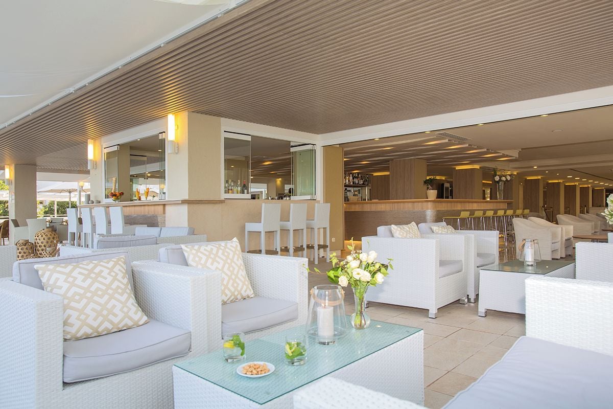 Ein moderner Außen-Loungesbereich mit weißen Korbmöbeln, einem Glastisch und einer Bar im Hintergrund.