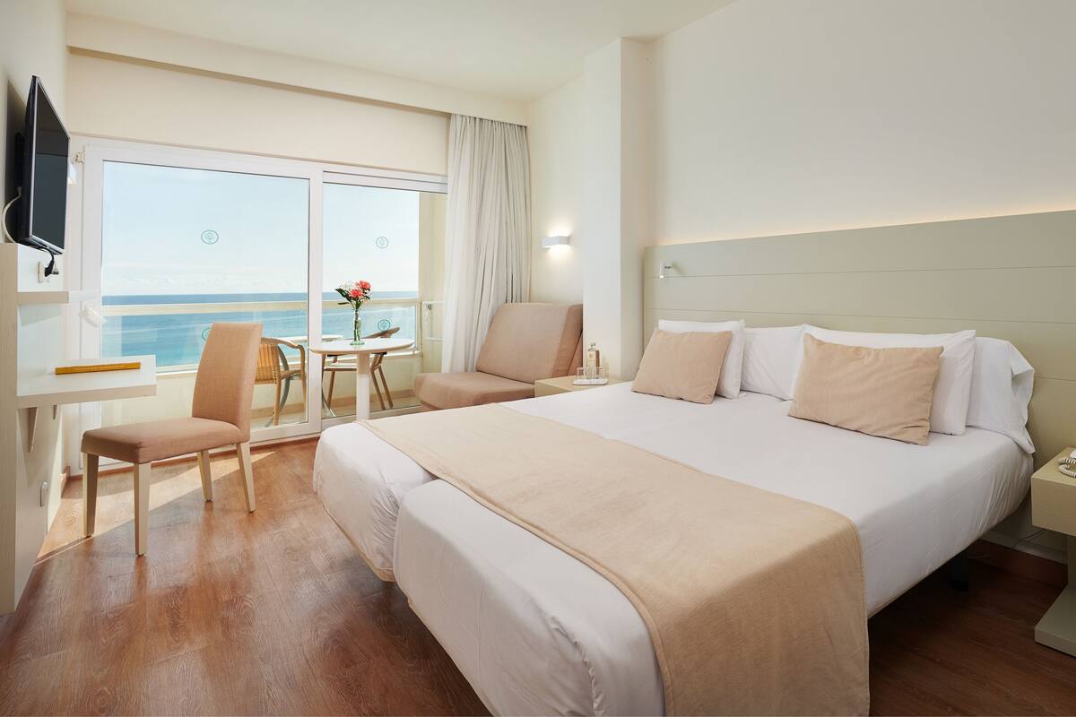 A hotel room with a bed, a desk, a chair, a small table with a vase, and a view of the ocean.