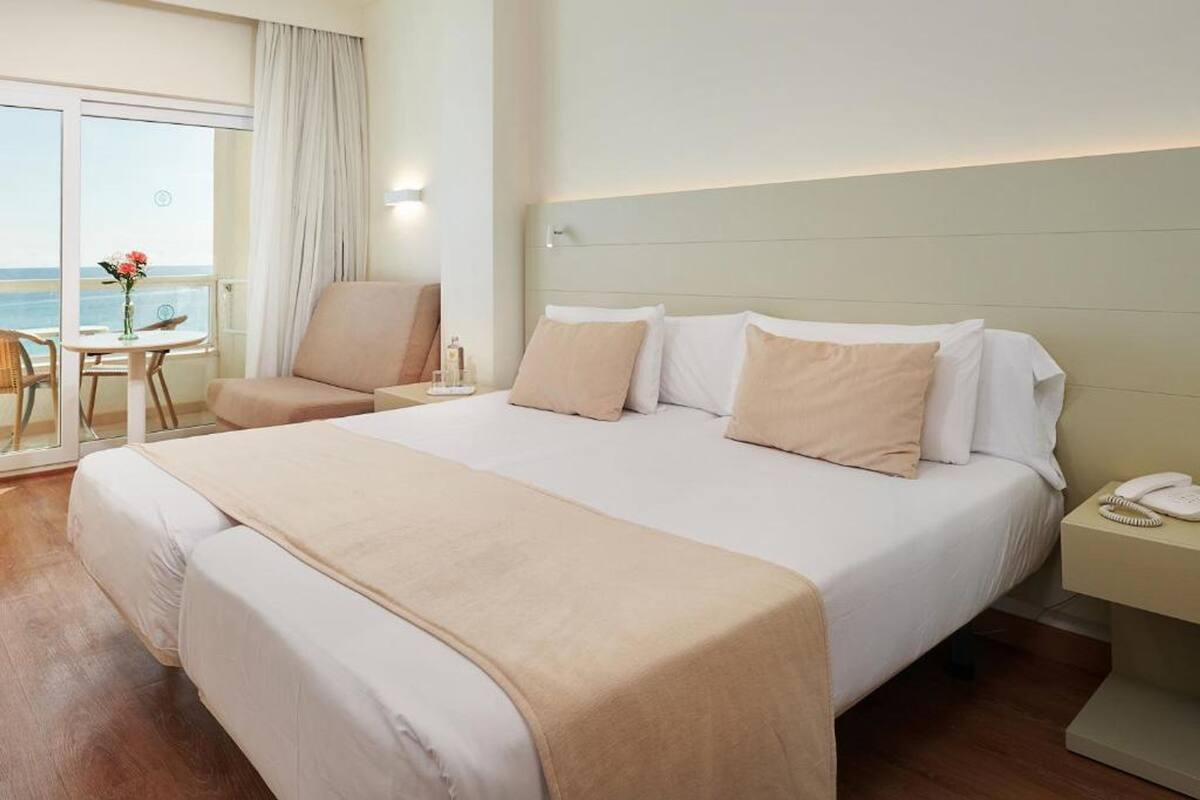 A hotel room with a large bed, a chair, a small table with a vase, and a view of the sea through a glass door.