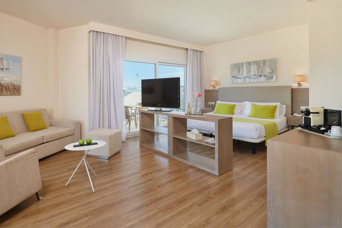 A hotel room with a sofa, a bed, a TV, a coffee machine, and a view of the sea.