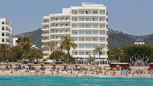A multi-story hotel with balconies, a beach in the foreground, and mountains in the background.