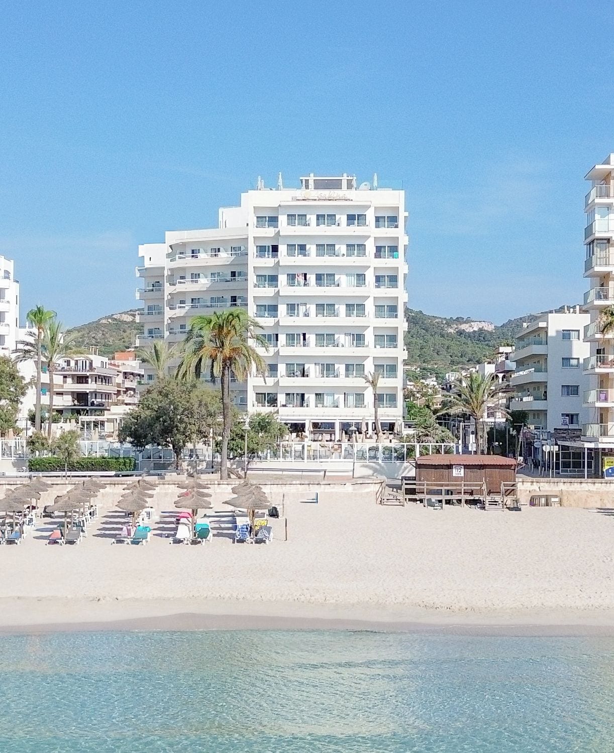 Ein Hotel direkt am Strand mit mehreren Etagen, ein Sandstrand im Vordergrund und ein klarer, blauer Himmel.