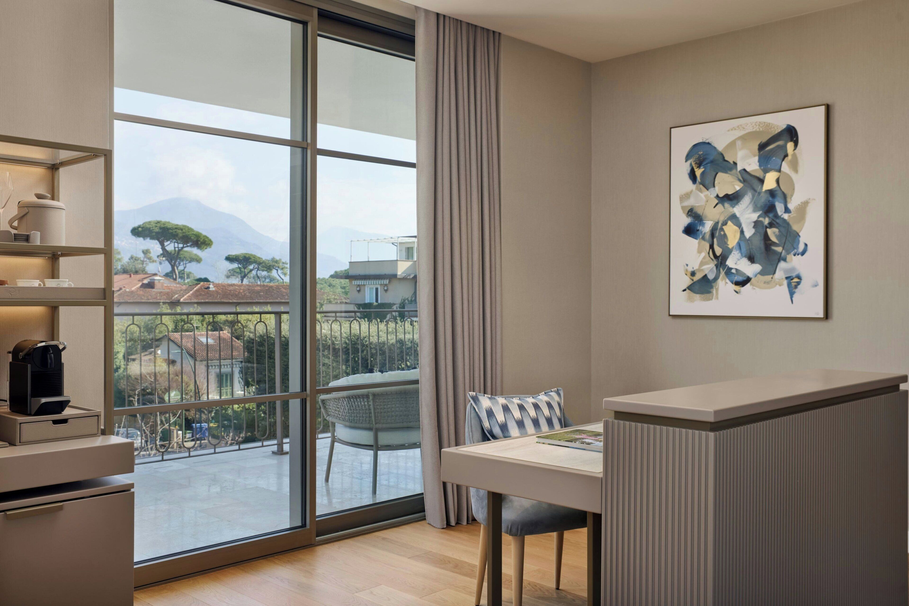 A modern hotel room with a large sliding glass door to a balcony, a dining area with a table and chairs, a coffee machine, and a framed abstract painting on the wall.