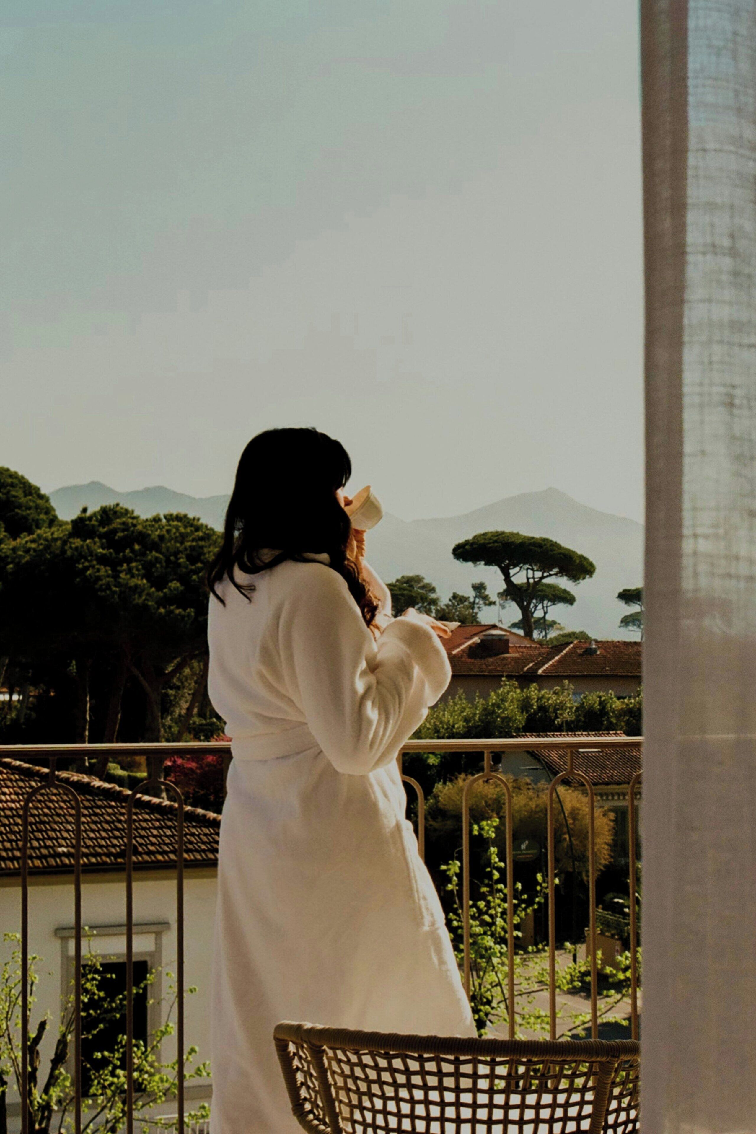 A person in a white robe standing on a balcony with a view of trees and mountains.