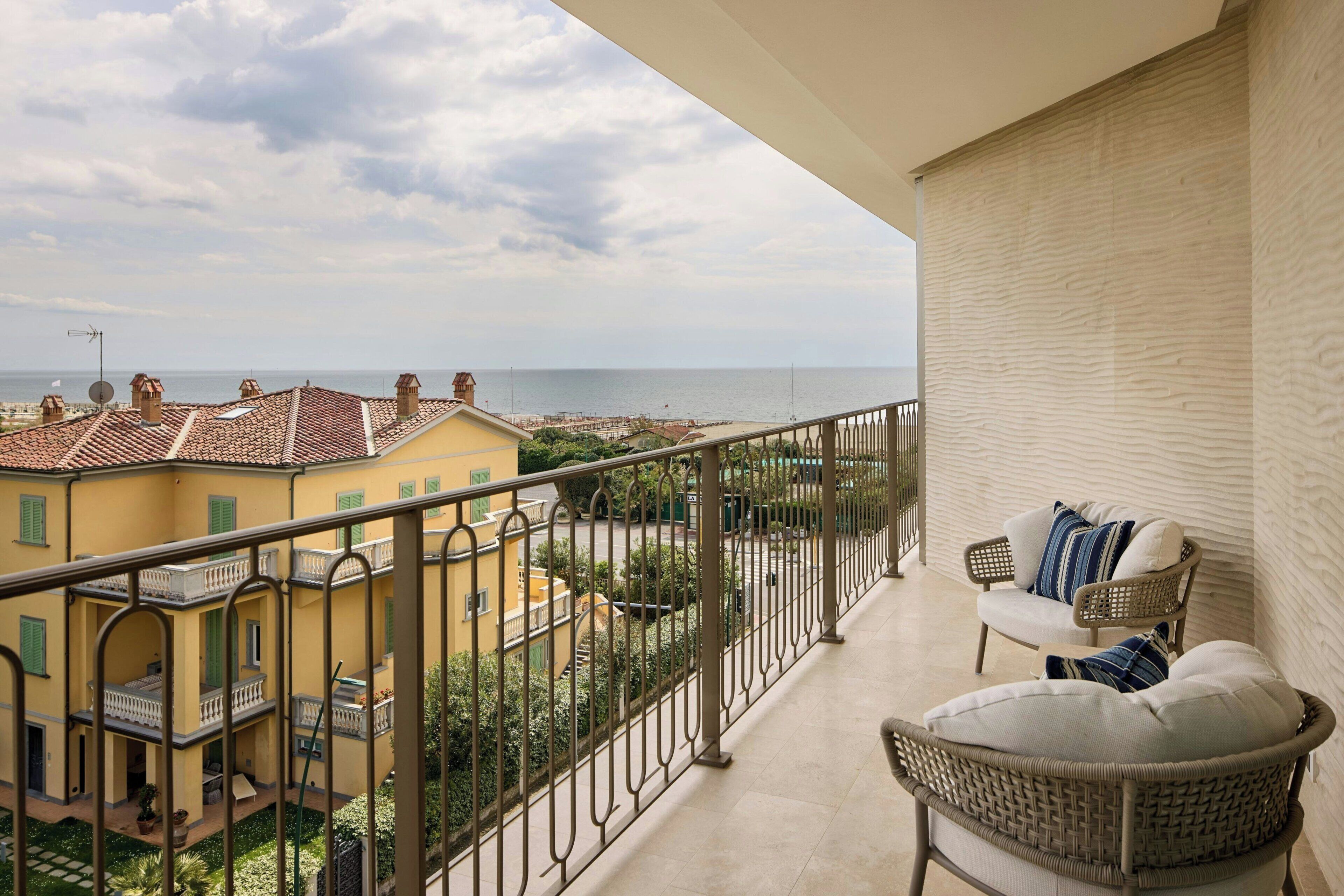 A balcony with a view of a coastal town, a wicker chair, and a railing.