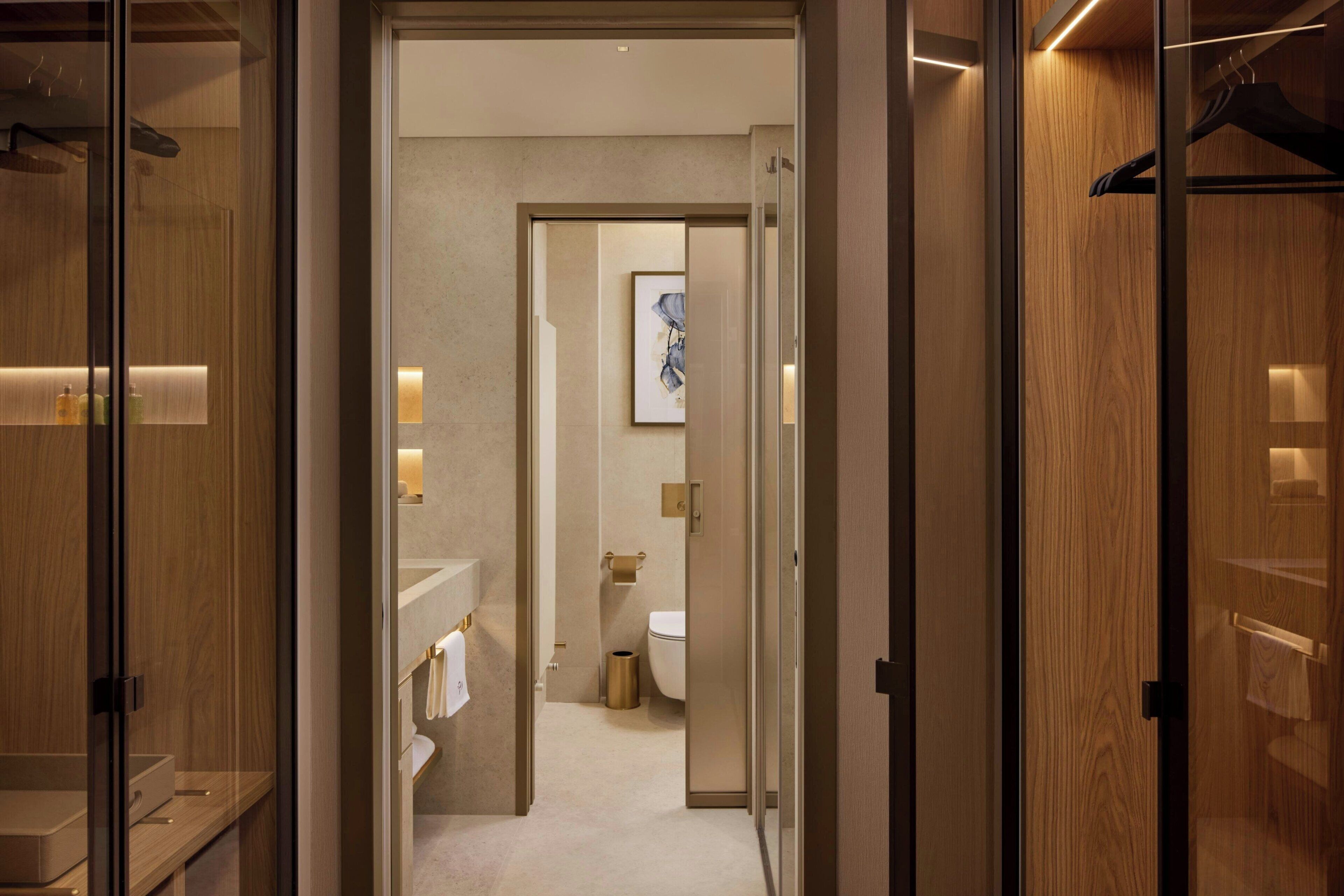 A hotel hallway leading to a bathroom with a toilet, sink, and towel rack.