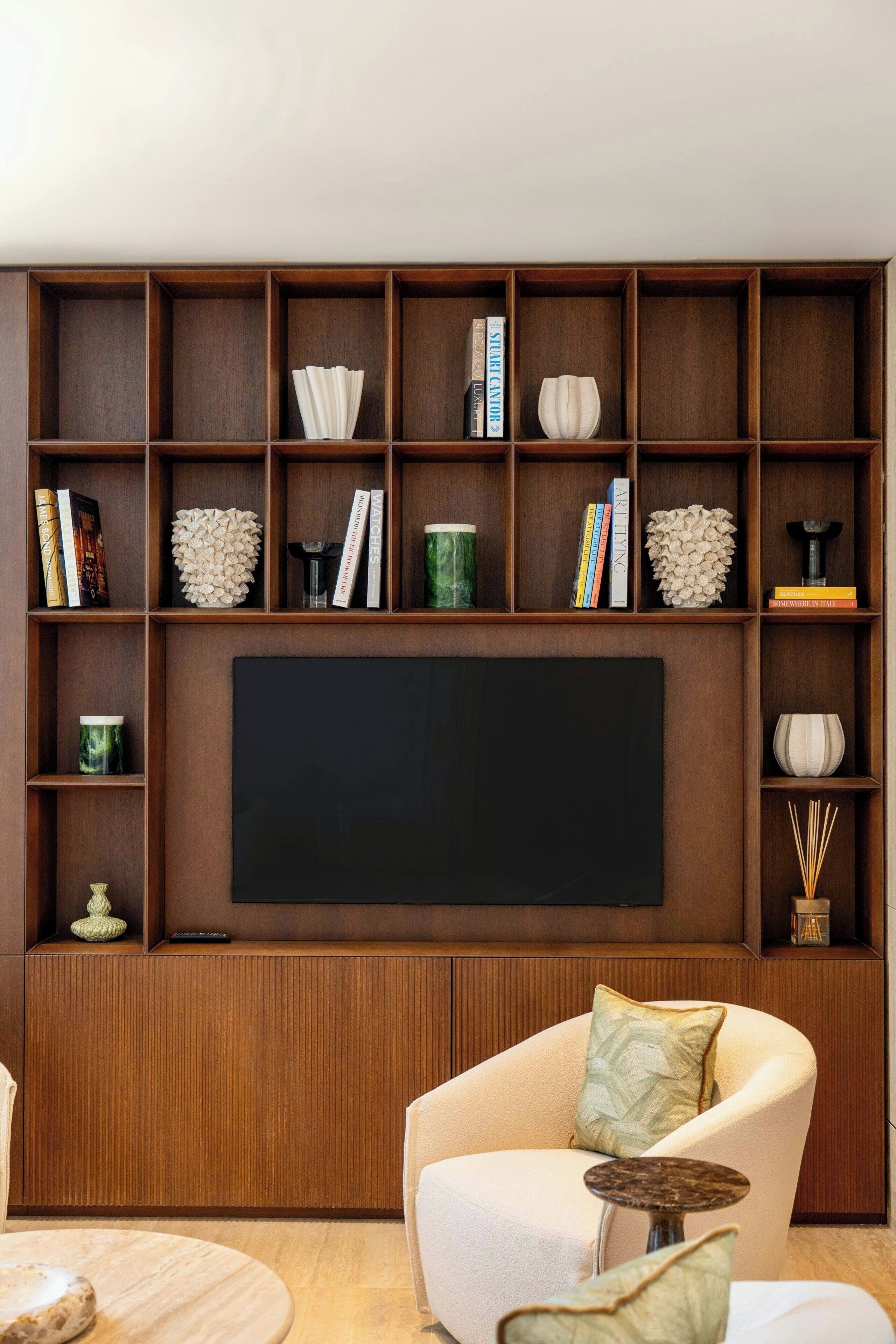 A living room with a large flat-screen TV, wooden shelving with books and decorative items, a white armchair, and a small round table.